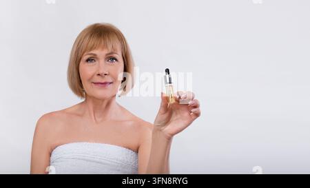 Smiling Middle-aged Caucasian Woman Holding A Serum Bottle With Dropper For Skincare Routine, Standing In Front Of Simple Background, Demonstrating Be Stock Photo