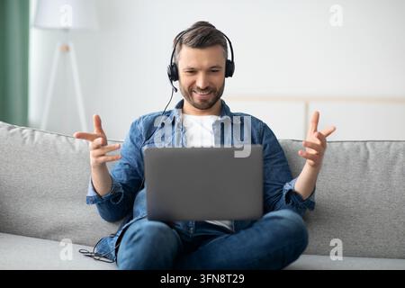 Positive bearded man having video conference from home, using headset and laptop, talking and gesturing. Cheerful middle-aged man resting on couch in Stock Photo
