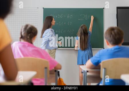 Schoolgirl solving mathematics problem on the chalkboard while her female teacher observing Stock Photo