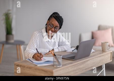 Cheerful african american business lady in formal outfit and eyeglasses attending webinar or business training, sitting at workdesk, using laptop at h Stock Photo