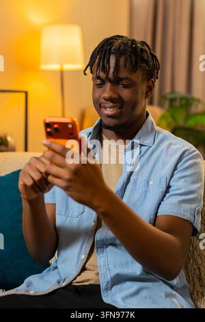 African American young man on home sofa checks morning notifications on smartphone with focused look and gentle smile. Black guy on home sofa reads me Stock Photo