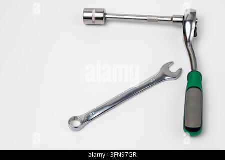 Chrome ratchet wrench with green and black handle rests on clean white surface next to combination wrench marked 10. Long extension bar connects to he Stock Photo