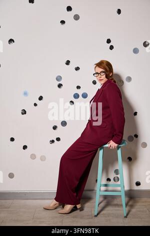 White redhead presenter leaning on stool rehearsing a talk in burgundy suit expressive gesture, polkadot backdrop, studio setting Stock Photo