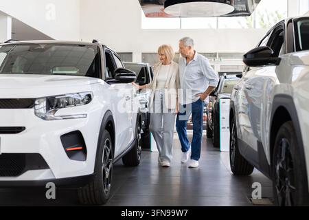 Senior Caucasian couple walking through modern car dealership showroom, comparing displayed vehicles Stock Photo