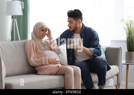 Aggressive arab man arguing with his upset pregnant muslim wife at home, sitting on couch in living room. Stressed expectant lady in hijab feeling unw Stock Photo