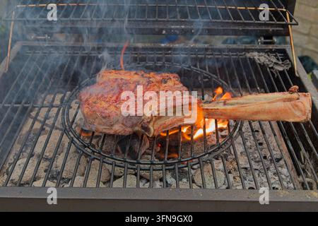 A thick, premium tomahawk ribeye steak searing over open flames and hot charcoal on an outdoor barbecue grill with smoke rising. Stock Photo