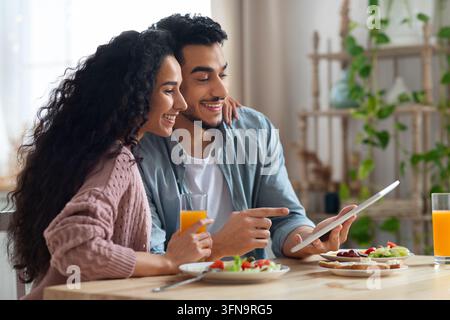 Young Arab Couple Using Digital Tablet In Kitchen While Having Breakfast. Cheerful Middle Eastern Spouses Enjoying Tasty Food And Watching Funny Video Stock Photo