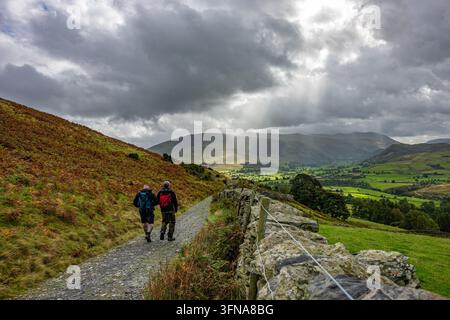 Hikers walk along a track by a drystone wall below sunbeams breaking through storm clouds over Lake District National Park, Cumbria, UK. Stock Photo