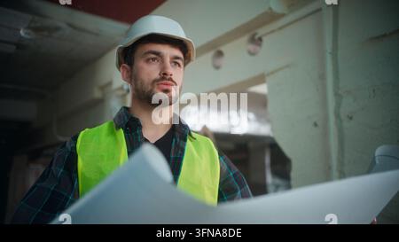 Serious Caucasian contractor man in uniform studying blueprint at construction site male architect builder in helmet holding draft engineer foreman Stock Photo