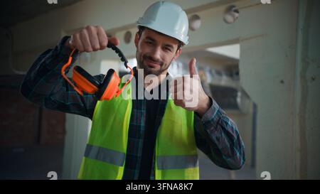 Cheerful Caucasian electrician man showing thumb up wearing safety earmuffs factory male guy technician builder engineer recommend acoustic headphones Stock Photo