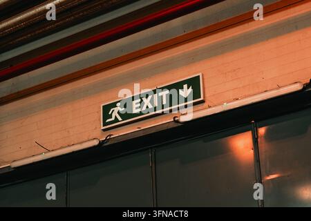 Illuminated green exit sign mounted on an indoor wall in an industrial setting. Concept of emergency evacuation, building safety compliance, wayfindin Stock Photo