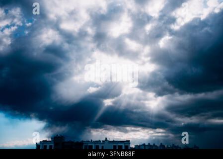 Dark storm clouds filling the sky, with bright sun rays piercing through, illuminating the city buildings below, symbolizing a challenging atmosphere transforming into hope Stock Photo