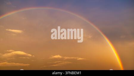 A full rainbow stretches across the sky above Thessaloniki, illuminated by warm sunset colors after rainfall, creating a spectacular weather phenomeno Stock Photo