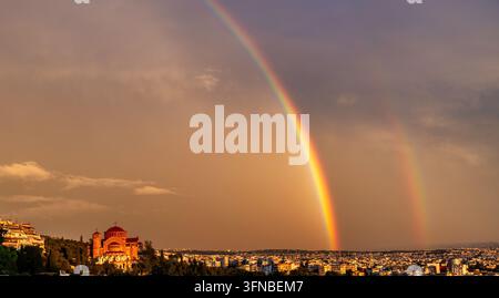 A dramatic double rainbow arches over Saint Paul Church and the urban landscape of Thessaloniki after rainfall, illuminated by warm sunset light and s Stock Photo