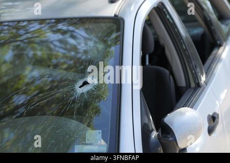 Car windshield with central bullet hole and radiating shattered safety glass, close up showing impact pattern and debris suggesting crime scene, vanda Stock Photo
