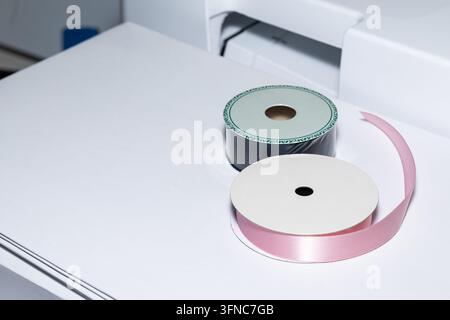 Pink satin ribbon and dark ribbon spools on white paper beside sewing machine, creating clean crafting Stock Photo