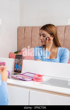 Young adult woman creating beauty content, applying makeup on her face, recording herself with a smartphone for a video tutorial or social media lives Stock Photo