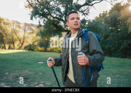 Male hiker with backpack and trekking poles walks through a sunlit green park, representing outdoor adventure, active travel, fitness, exploration, an Stock Photo