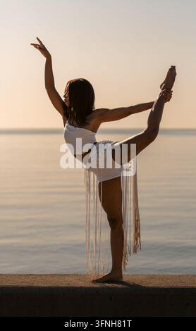 A beautiful Cuban woman performs the standing "scorpion" dance pose in the warm light of the golden hour, backlit; she wears a white one-shoulder crop Stock Photo