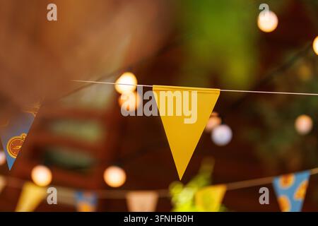 Selective focus shot of a yellow triangular festival flag hanging on a line with glowing bokeh background lights. Stock Photo