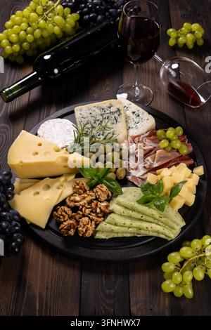 Charcuterie platter presenting various cheeses, prosciutto, olives, nuts, and crackers, accompanied by red wine bottle, glasses, and fresh grapes on a Stock Photo