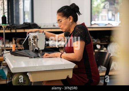 Skilled female artisan repairs a dark garment on an industrial sewing machine in a textile workshop, concentrating at her worktable among fabric and t Stock Photo