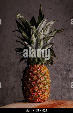 Studio still life of a ripe pineapple standing upright on a rustic wooden table, set against a dark textured grunge wall. Tropical fruit concept with Stock Photo