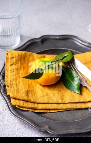 Fresh mandarin citrus with green leaves rests on a folded mustard linen napkin, arranged on a dark metal tray beside a glass and silver cutlery. Stock Photo