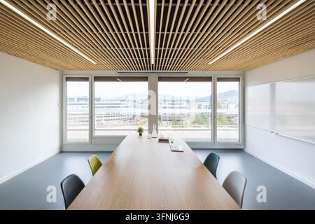 Bright modern coworking office featuring a long conference table, colorful chairs, whiteboard, wooden slat ceiling, and panoramic windows overlooking Stock Photo