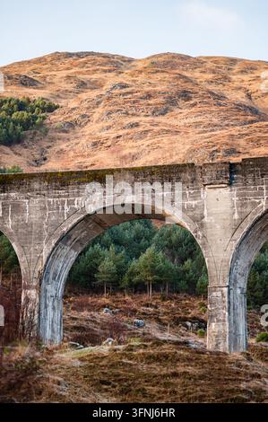 Weathered stone viaduct with broad arches spans a wooded valley beneath dry, rugged hills, showcasing historic engineering and a dramatic rural landsc Stock Photo