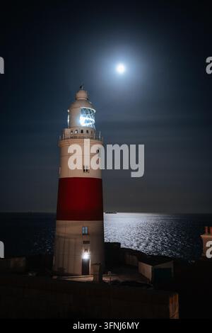 Historic lighthouse in Gibraltar shines beside the moonlit sea at night, with a bright full moon reflecting on calm water beneath a dark, dramatic sky Stock Photo