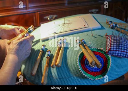 Close-up shot showing the hands of a senior woman working with wooden bobbins and threads on a lace pillow, capturing the intricate details of traditi Stock Photo