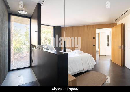 A bright, contemporary bedroom in a luxury villa in Catalonia, Spain, featuring a black shower, a white bed, wood paneling, large windows, and a sleek Stock Photo