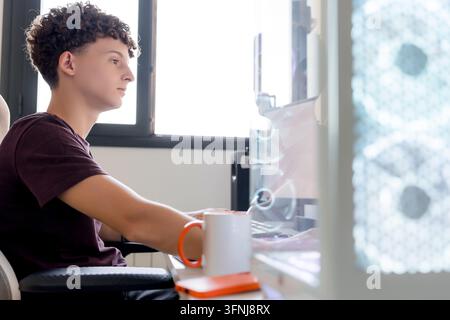Young man works at a desktop gaming computer in a home office, using AI tools while seated at a desk beside a mug and smartphone in daylight. Stock Photo