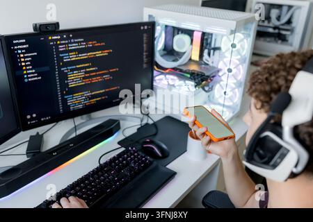 Young man wearing virtual reality headset uses a smartphone beside a gaming computer with code editor on monitor, illuminated keyboard and desktop PC Stock Photo