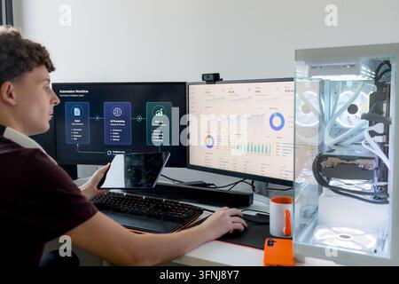 Young man works with AI tools and analytics dashboards on dual monitors, using a tablet beside a custom gaming computer in a modern home office. Stock Photo