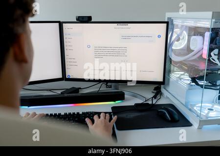Young man uses an AI assistant on a dual-monitor gaming computer setup with RGB keyboard, webcam and illuminated desktop tower in a modern home worksp Stock Photo