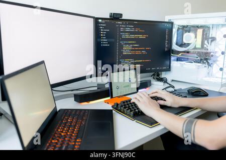 Close-up of a young man using AI tools to write code on a gaming computer, with multiple monitors, laptop, tablet, mechanical keyboard, and illuminate Stock Photo