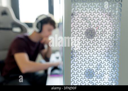 Selective focus on a gaming computer case while a young man wearing headphones uses AI tools at a desk, illustrating technology, gaming, and digital c Stock Photo