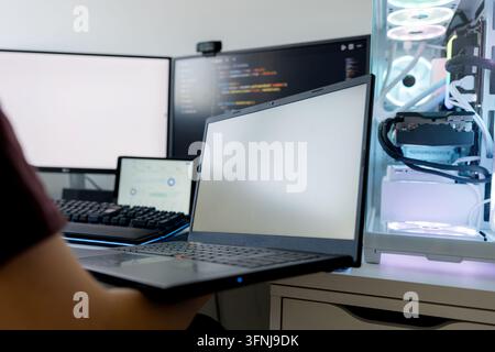 Young man works with AI tools on a laptop beside dual monitors and a custom gaming computer, creating code at a modern home technology workstation. Stock Photo