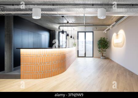 Bright modern coworking office interior featuring a curved orange brick reception desk, wood floor, exposed ceiling ducts, pendant lights, large windo Stock Photo
