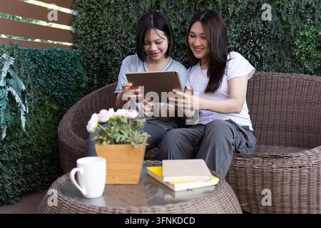 Shared Moment: Two friends, engrossed in the digital world, share a moment of connection while relaxing on a cozy patio, enhanced by natural greenery Stock Photo