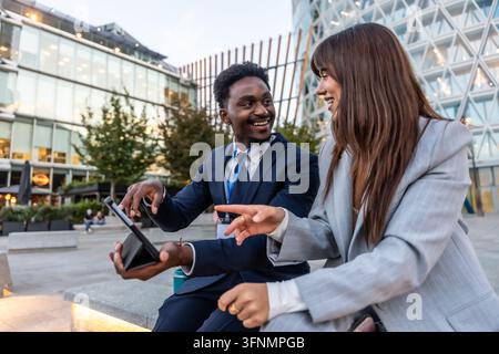 Two diverse business professionals collaborating and discussing information on a digital tablet, sitting on an urban bench outside modern office build Stock Photo