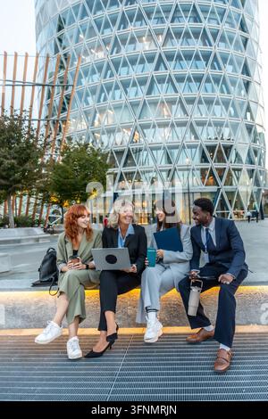 Diverse business professionals networking and collaborating outdoors in a modern urban setting, smiling and engaging in conversation while using a lap Stock Photo