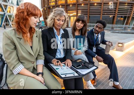 Diverse business professionals collaborating on strategy and analyzing financial data together during an informal outdoor meeting, fostering mentorshi Stock Photo