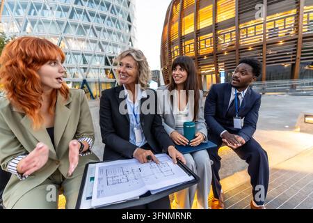 Diverse business professionals reviewing architectural blueprints and collaborating on a project, sitting outdoors in front of modern corporate buildi Stock Photo