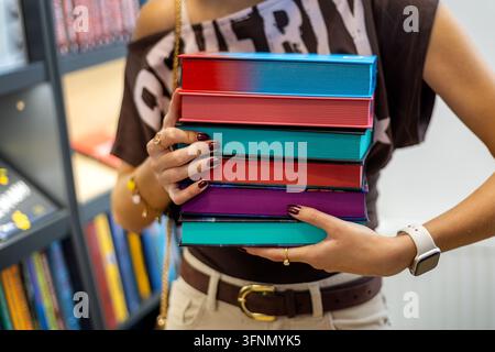 Woman carrying a stack of colorful hardcover books with bright painted page edges, modern bookstore scene representing reading and education. Stock Photo