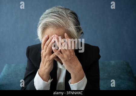 Overworked tired old lady holding head feeling headache. Asian senior business woman stress from hard work. Frustrated business woman thinks seated at Stock Photo