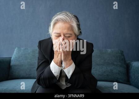 Overworked tired old lady holding head feeling headache. Asian senior business woman stress from hard work. Frustrated business woman thinks seated at Stock Photo