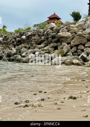 Shallow waves reach a rocky beach beneath a distant pavilion in soft daylight. Stock Photo